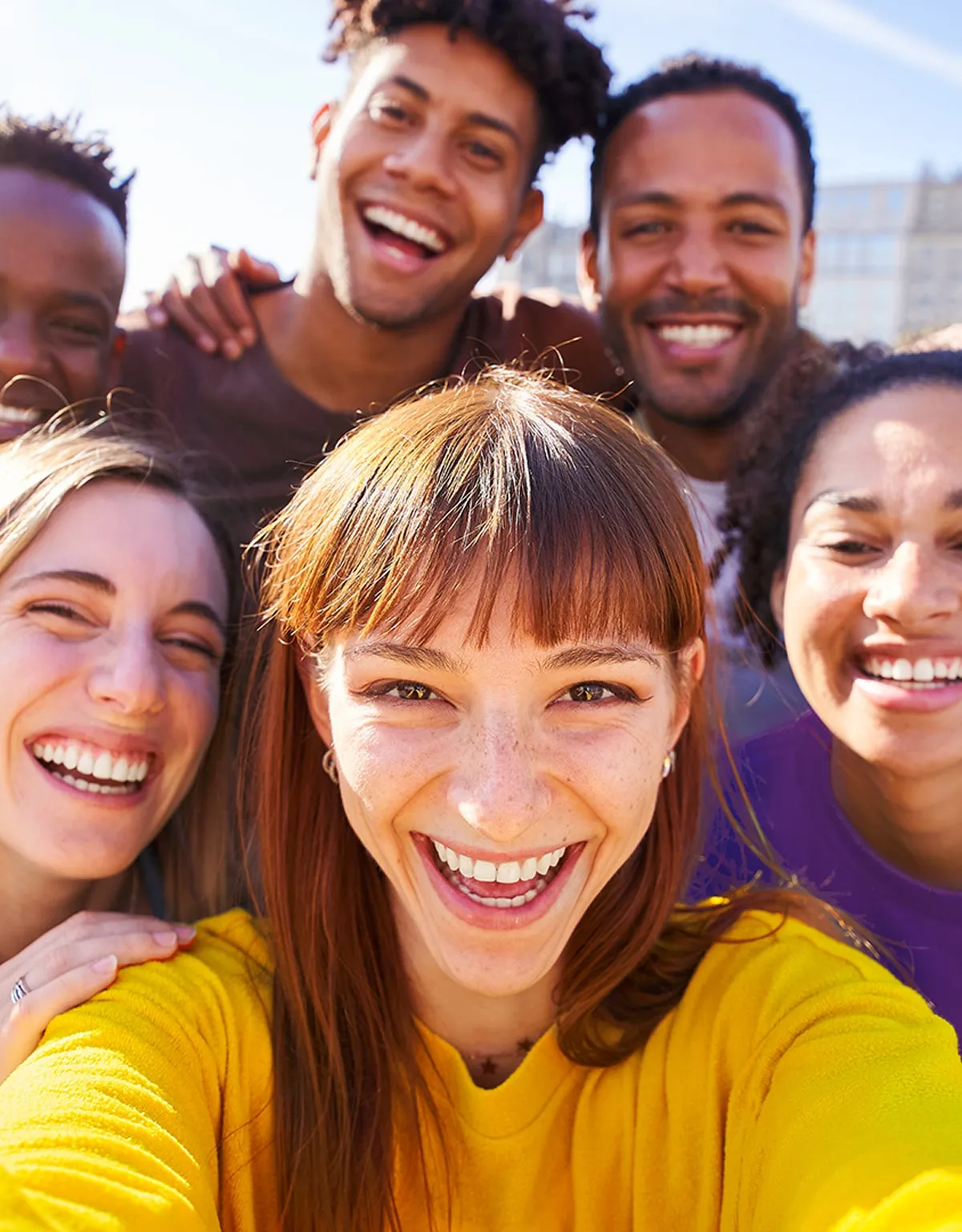 Group of six diverse friends smiling and taking a cheerful selfie outdoors on a sunny day.