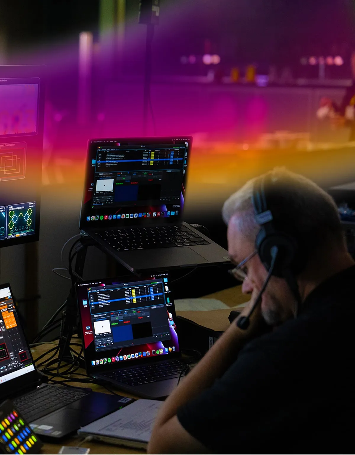 Man wearing headset working in a dimly lit room with multiple laptops and monitors displaying audio and video editing software.