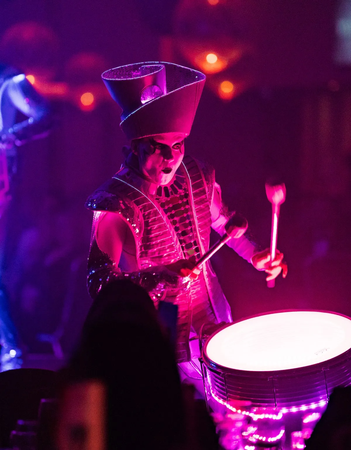 Performer in elaborate costume and tall hat playing a brightly lit drum with drumsticks in a dark, colorful stage setting.