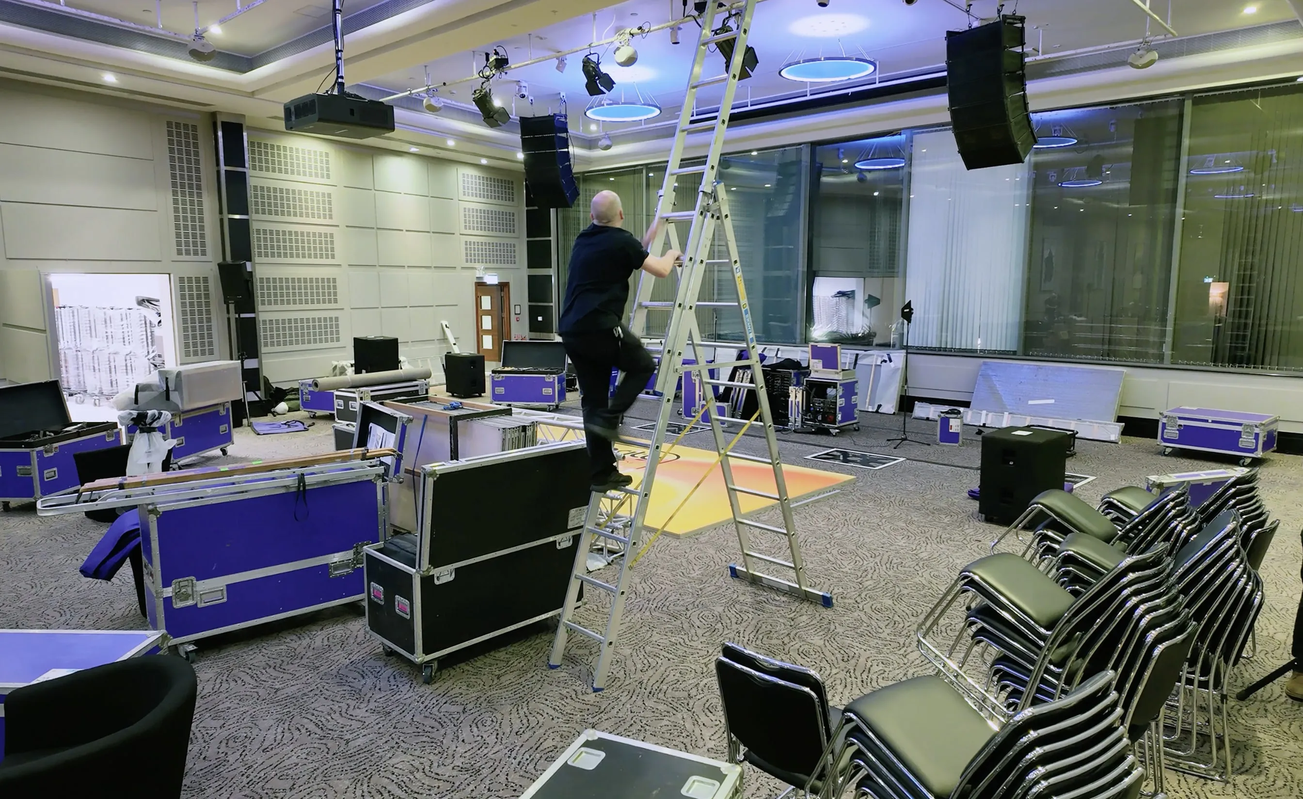 Man climbing a ladder in a large room filled with stacked chairs and equipment cases.