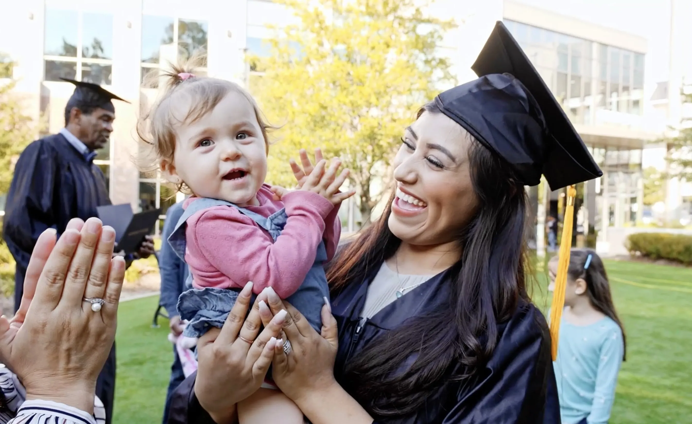 Smiling female graduate in cap and gown holding a clapping baby outside on a sunny day.