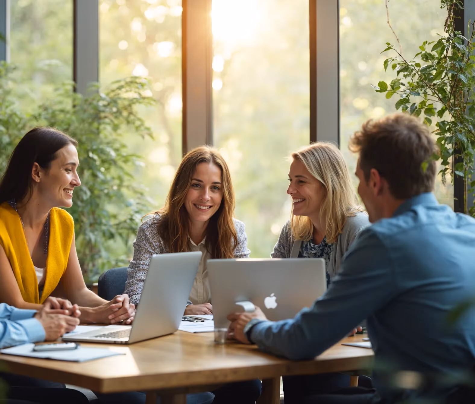 Four colleagues smiling and discussing work around a table with laptops and documents in a bright office with large windows and plants.