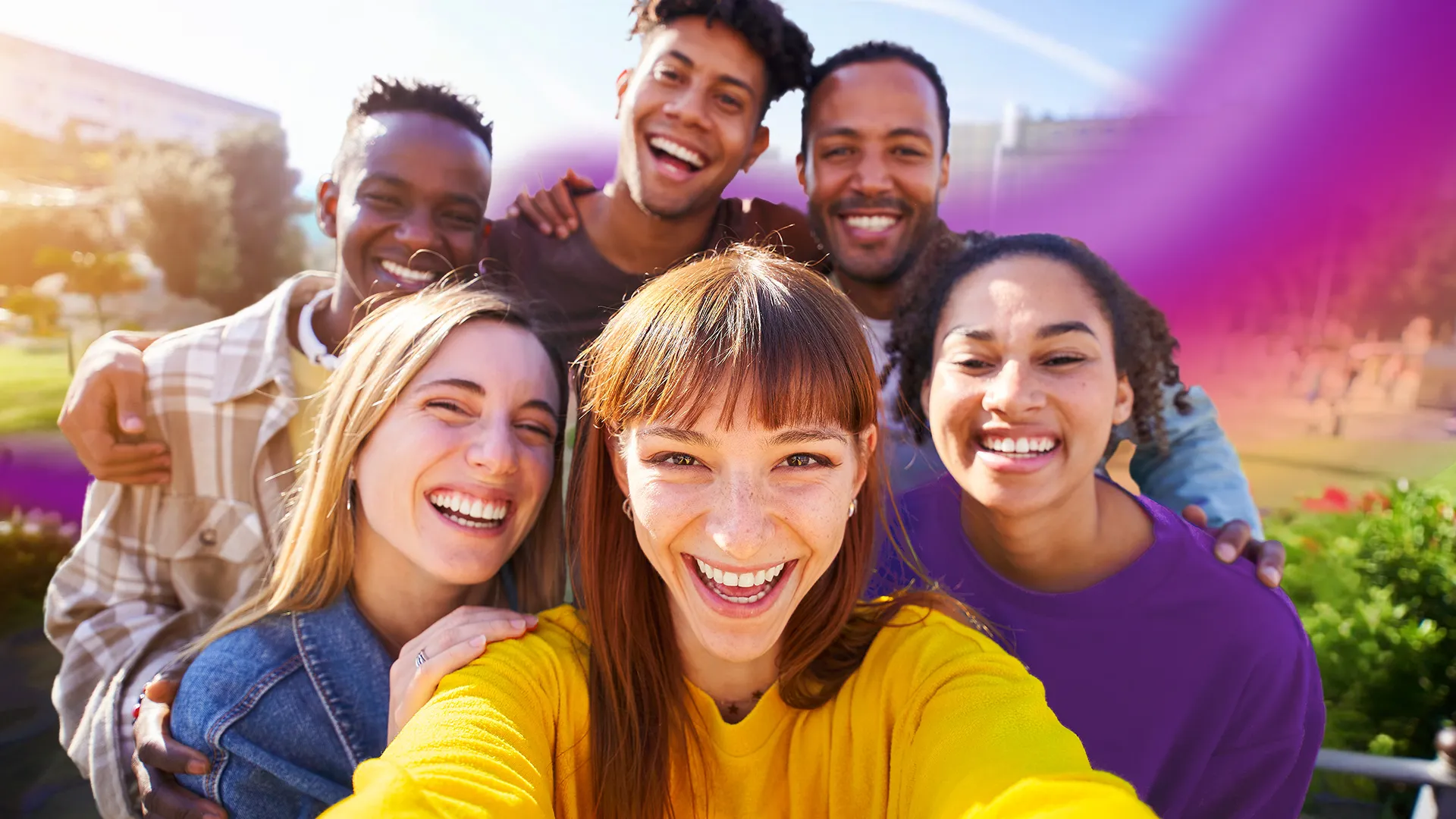 Six diverse young adults smiling and taking a selfie outdoors in a sunny park.