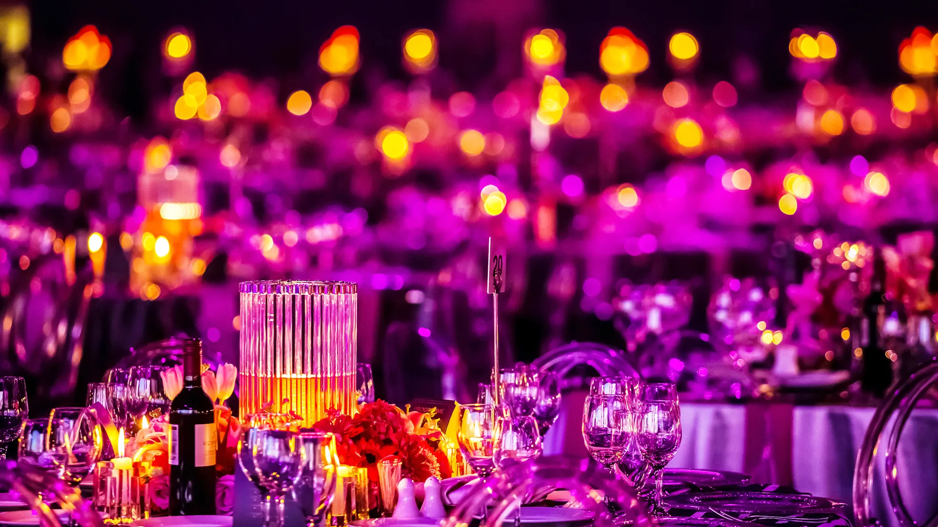 Elegant banquet tables decorated with candles, wine bottles, crystal glassware, and floral centerpieces under purple and warm lighting.