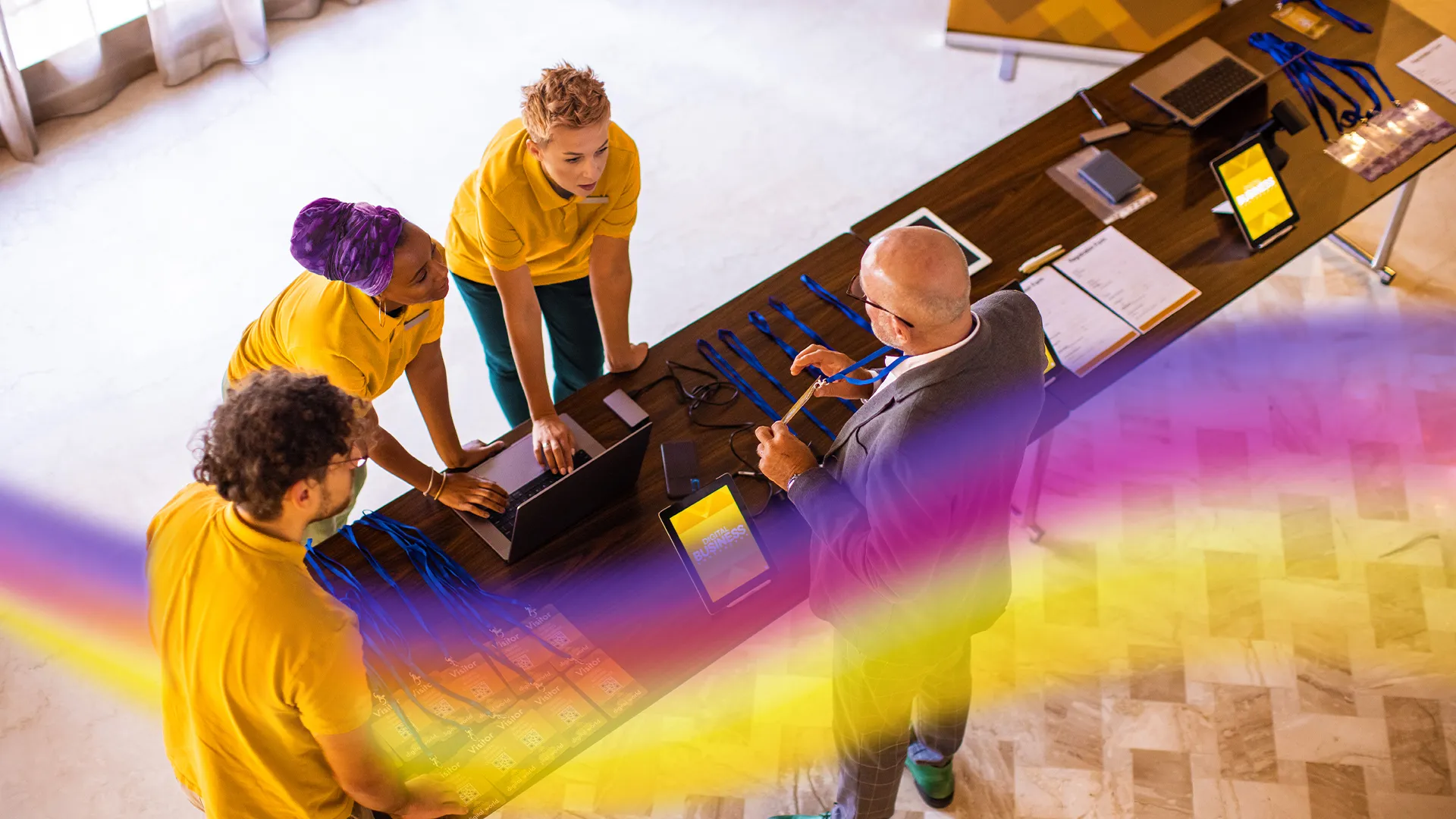 Three event staff in yellow shirts assisting a bald man with visitor badges at a registration desk.