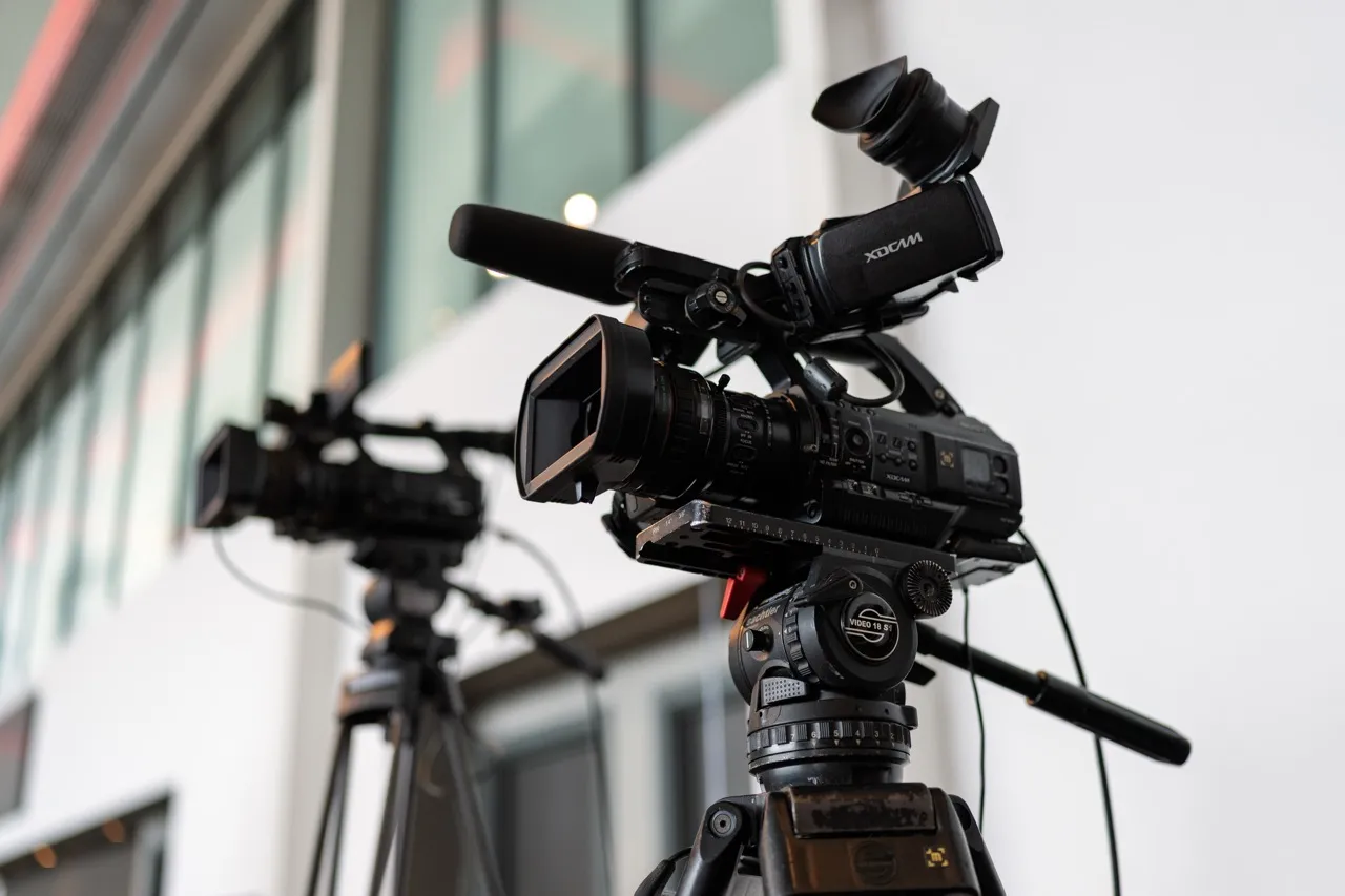 Two professional video cameras on tripods set up indoors with blurred windows in the background.