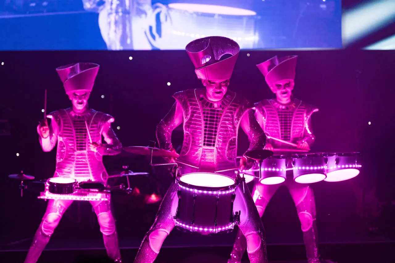 Three performers in illuminated futuristic costumes playing drums with vibrant purple and pink lighting.