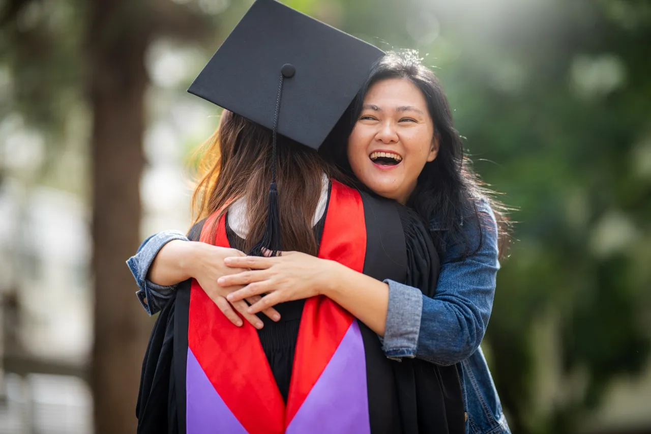 Smiling woman in a denim jacket hugging a graduate wearing a black cap and gown with red and purple stole outdoors.