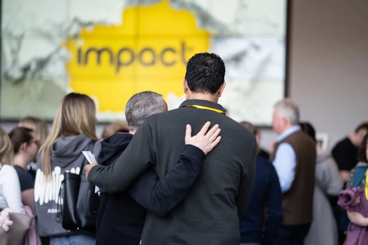 Two people with arms around each other at a conference with a blurred screen displaying the word 'impact' in the background.