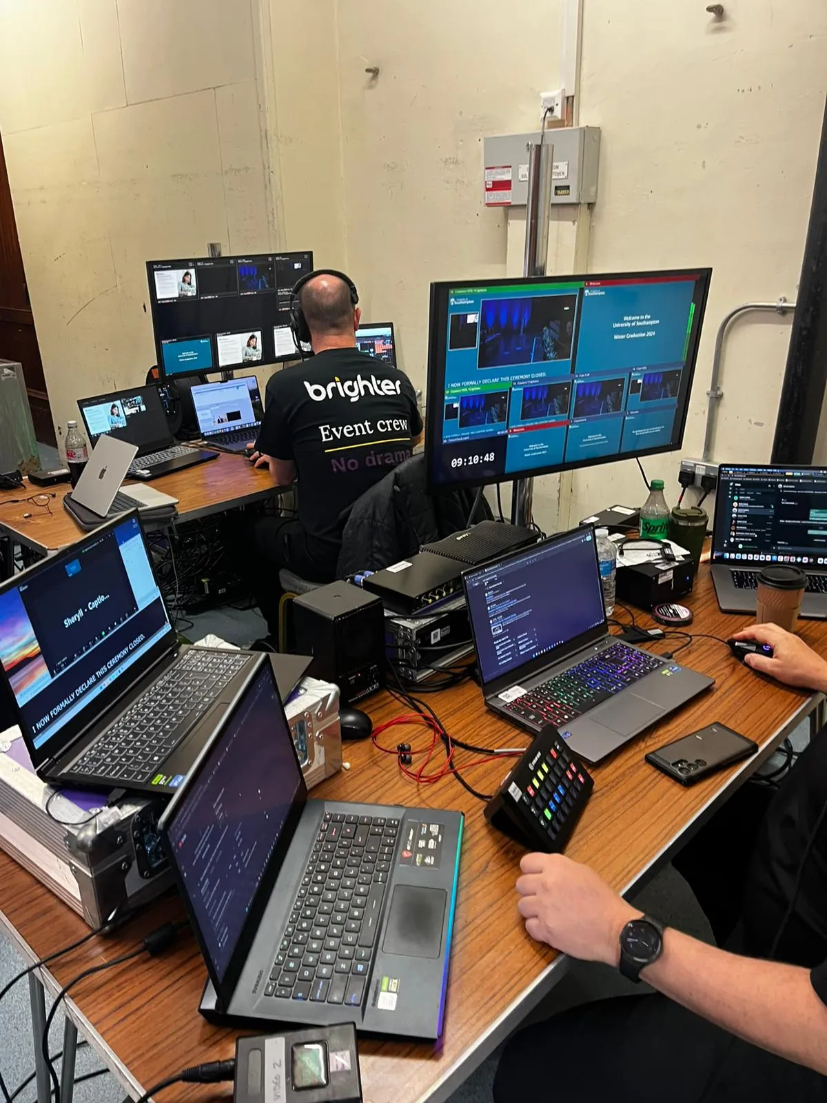 Two people operating multiple laptops and large monitors in a control room for a live event, one wearing a 'brighter Event crew No drama' shirt.