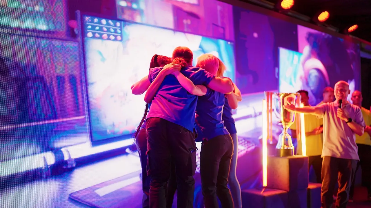 Group of esports players in blue shirts embracing in a team hug on stage with a trophy and announcer in the background.