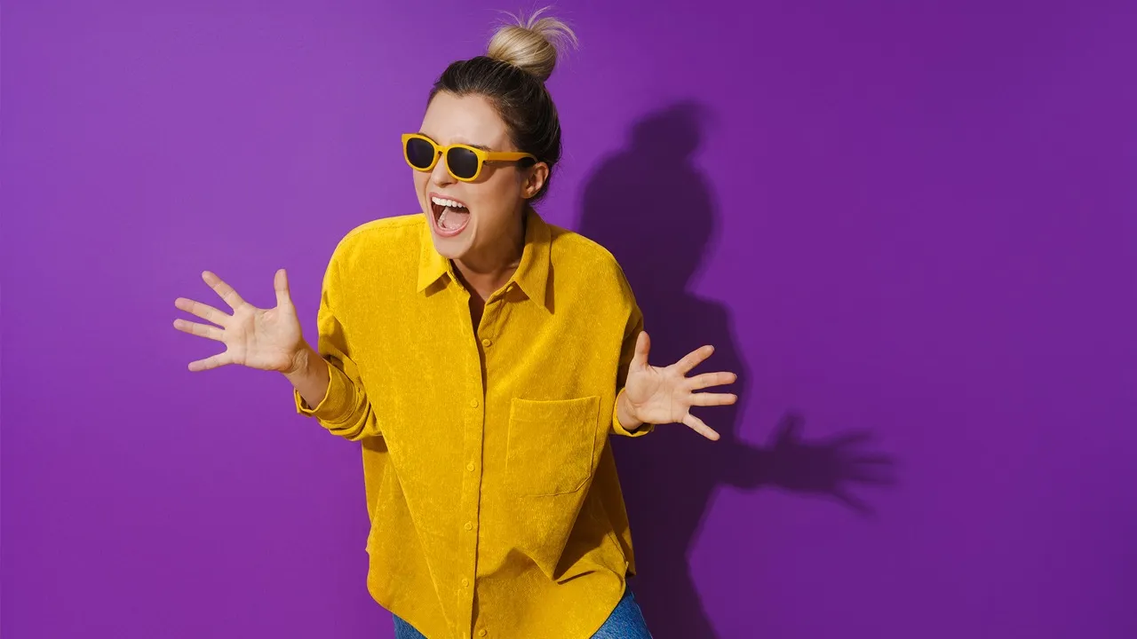 Smiling woman wearing yellow shirt and sunglasses against a purple background, expressing excitement with hands spread wide.