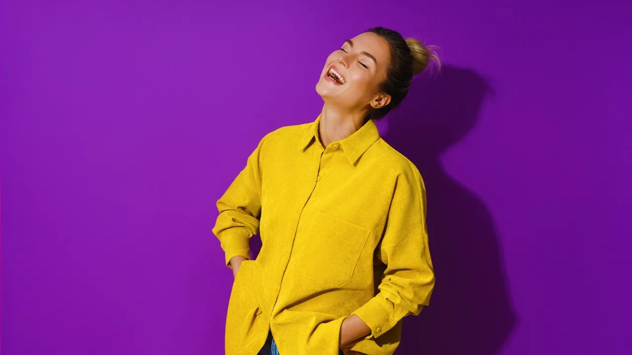 Smiling woman wearing a bright yellow jacket standing against a purple background with hands in pockets.