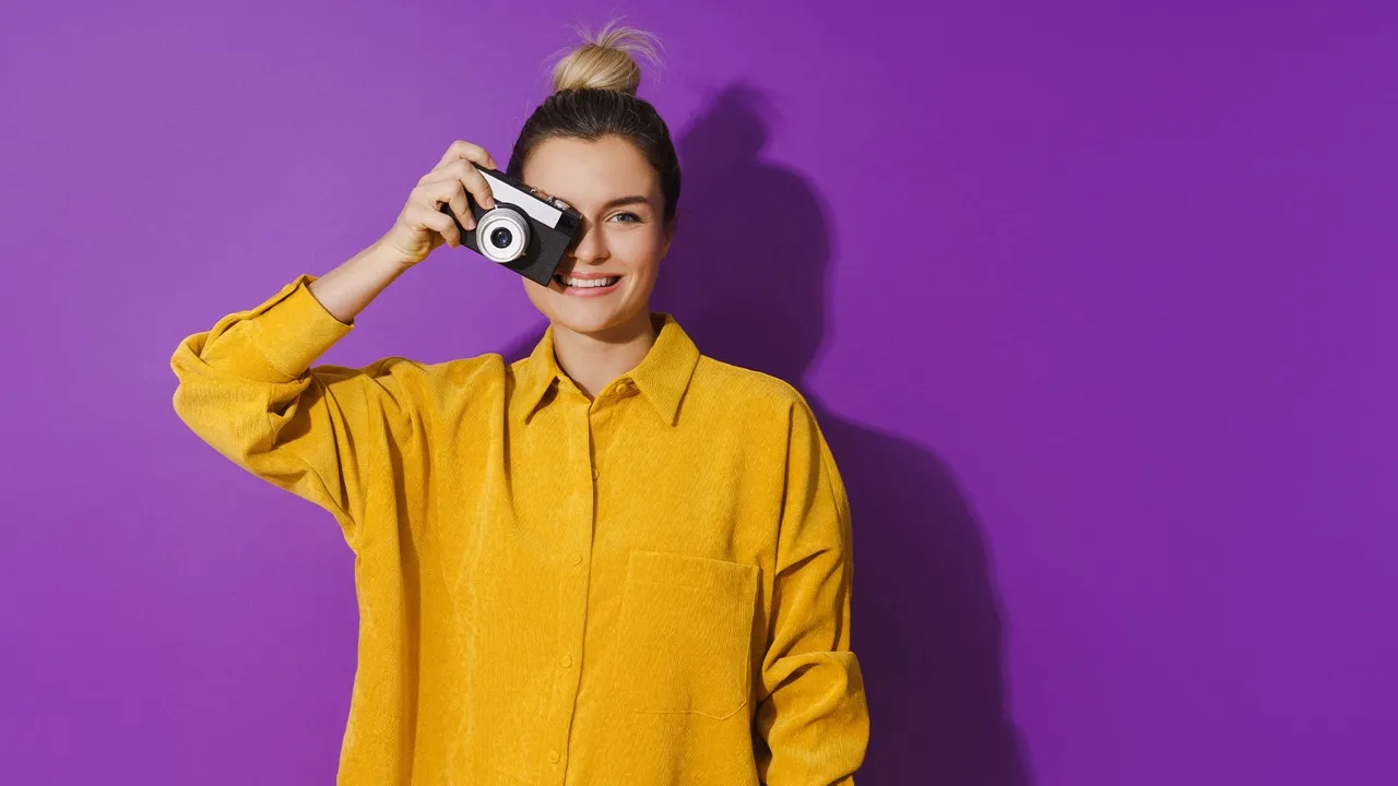Smiling woman in a yellow shirt holding a camera up to one eye against a purple background.