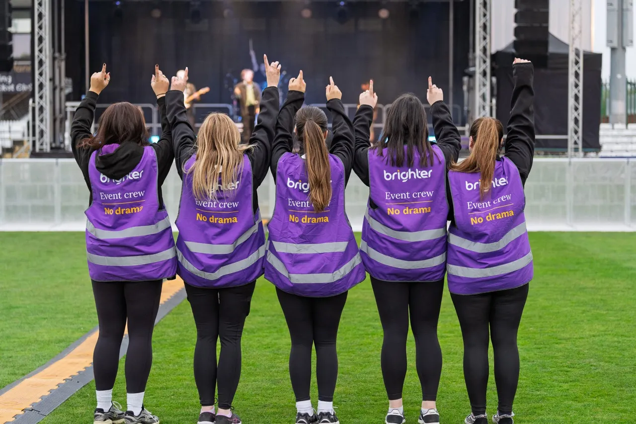 Five event crew members wearing purple 'No drama' vests with raised fingers facing a concert stage.