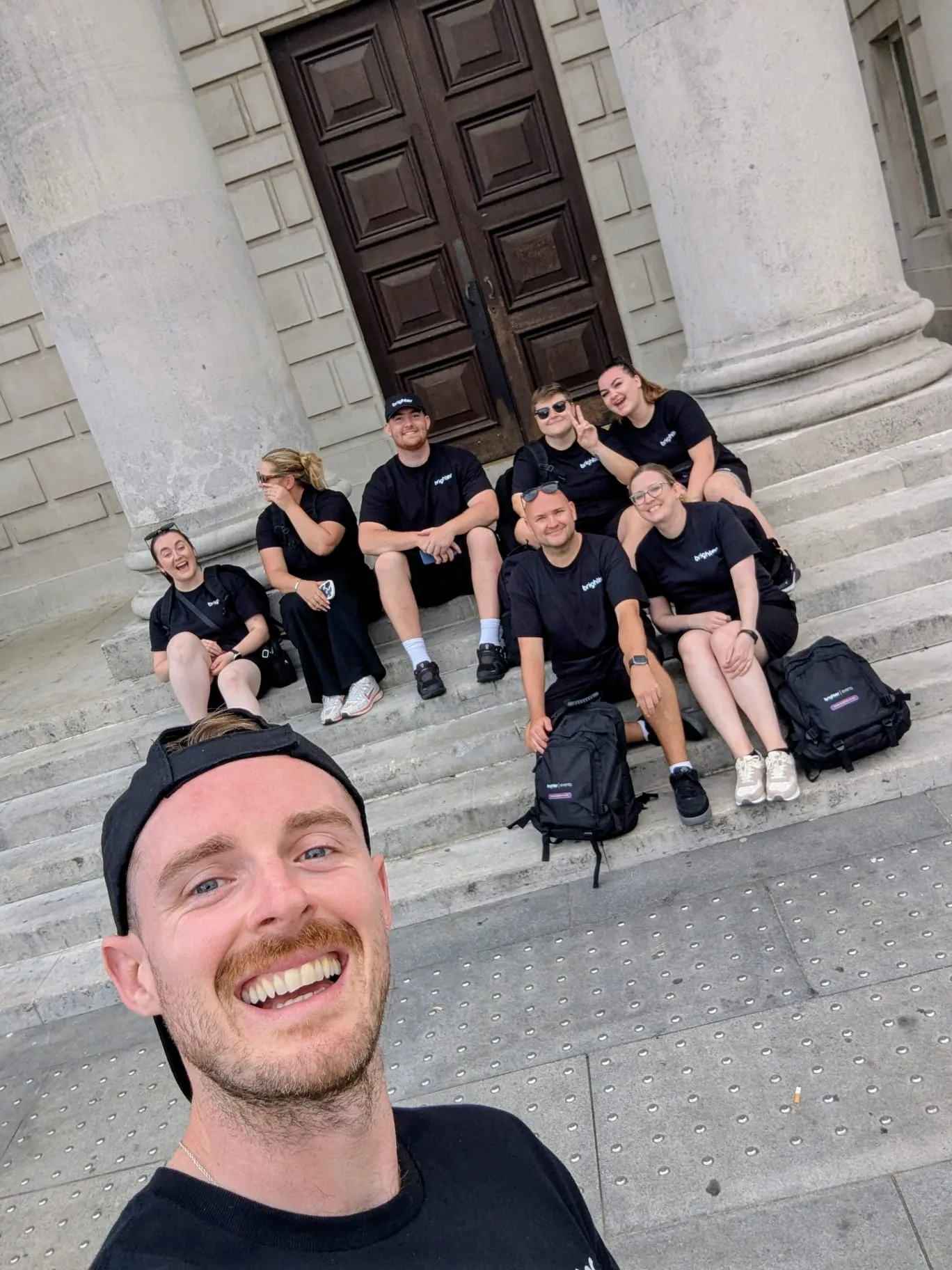 Group of seven adults in black shirts sitting on stone steps in front of a large wooden door, with one person in the foreground taking a smiling selfie.