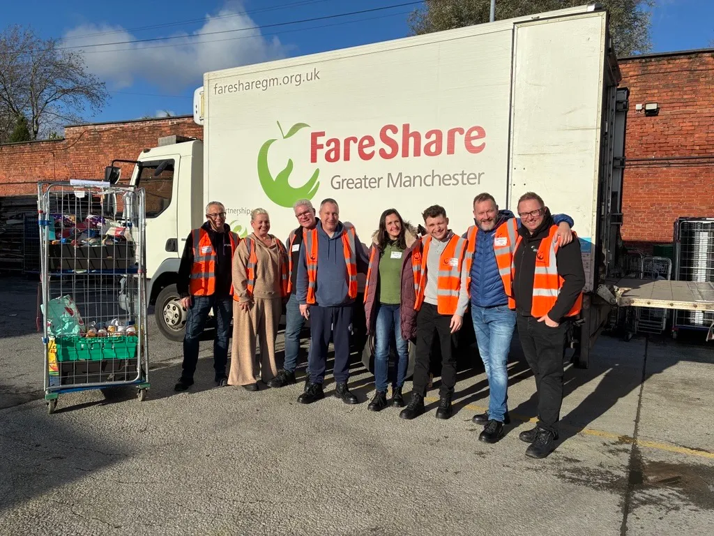 Group of seven people wearing high-visibility orange vests standing in front of a FareShare Greater Manchester delivery truck with a cart of food items beside them.