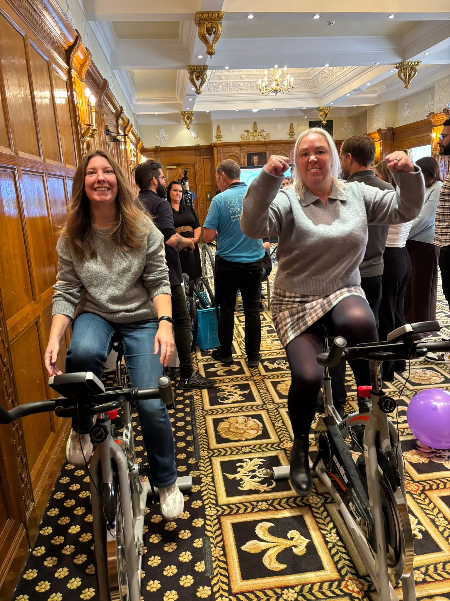 Two women smiling and posing on stationary bikes in a decorated indoor room with people standing in the background.