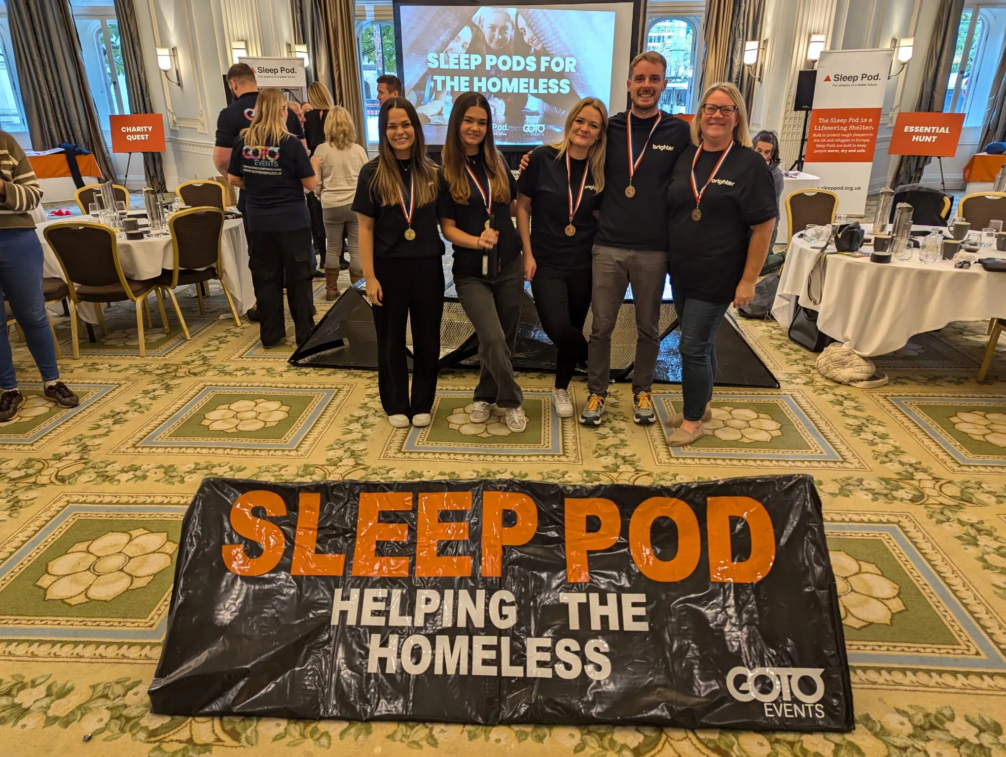 Five people wearing medals stand behind a Sleep Pod banner that says 'Helping the Homeless' at an indoor event.