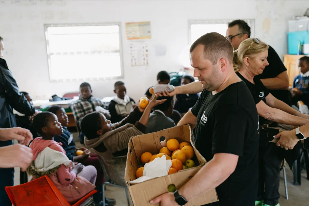 Volunteers distributing oranges and snacks to children seated inside a bright room.