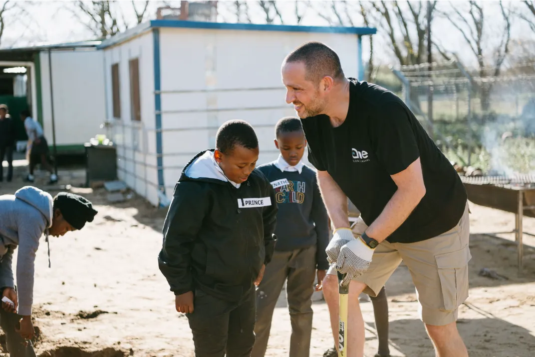 Man wearing gloves and a black t-shirt engaging with two young boys outdoors near a building on a sunny day.