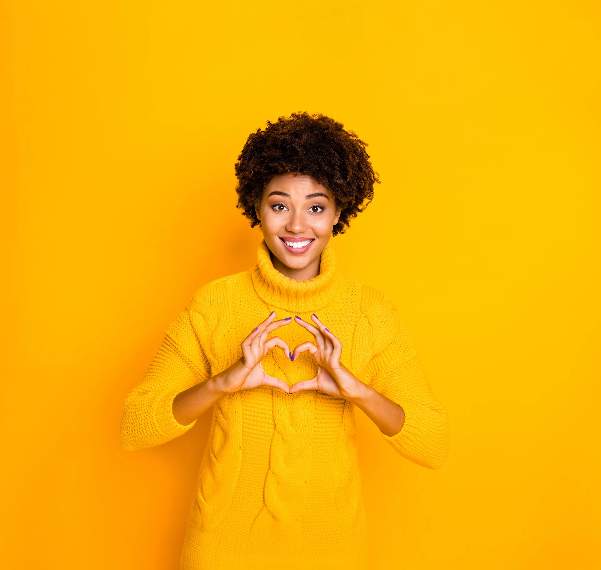 Smiling woman in a yellow sweater making a heart shape with her hands against a yellow background.