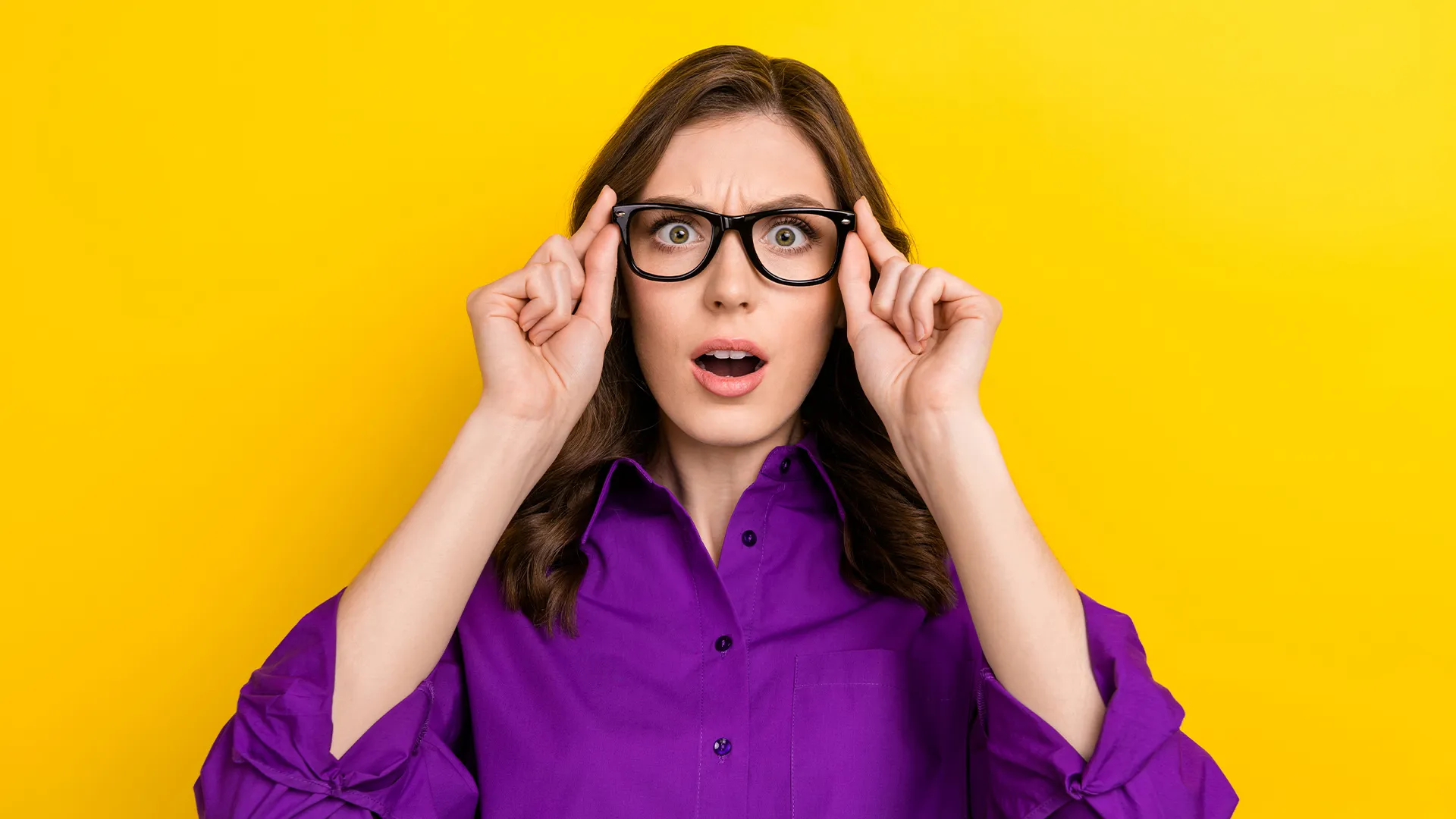 Woman in a purple shirt adjusting her black glasses with a surprised expression on a yellow background.