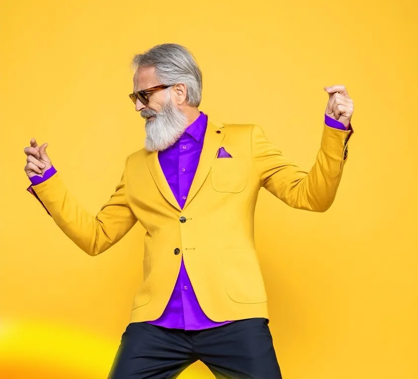 Stylish older man with gray hair and beard wearing sunglasses, a yellow blazer, and a purple shirt dancing against a yellow background.