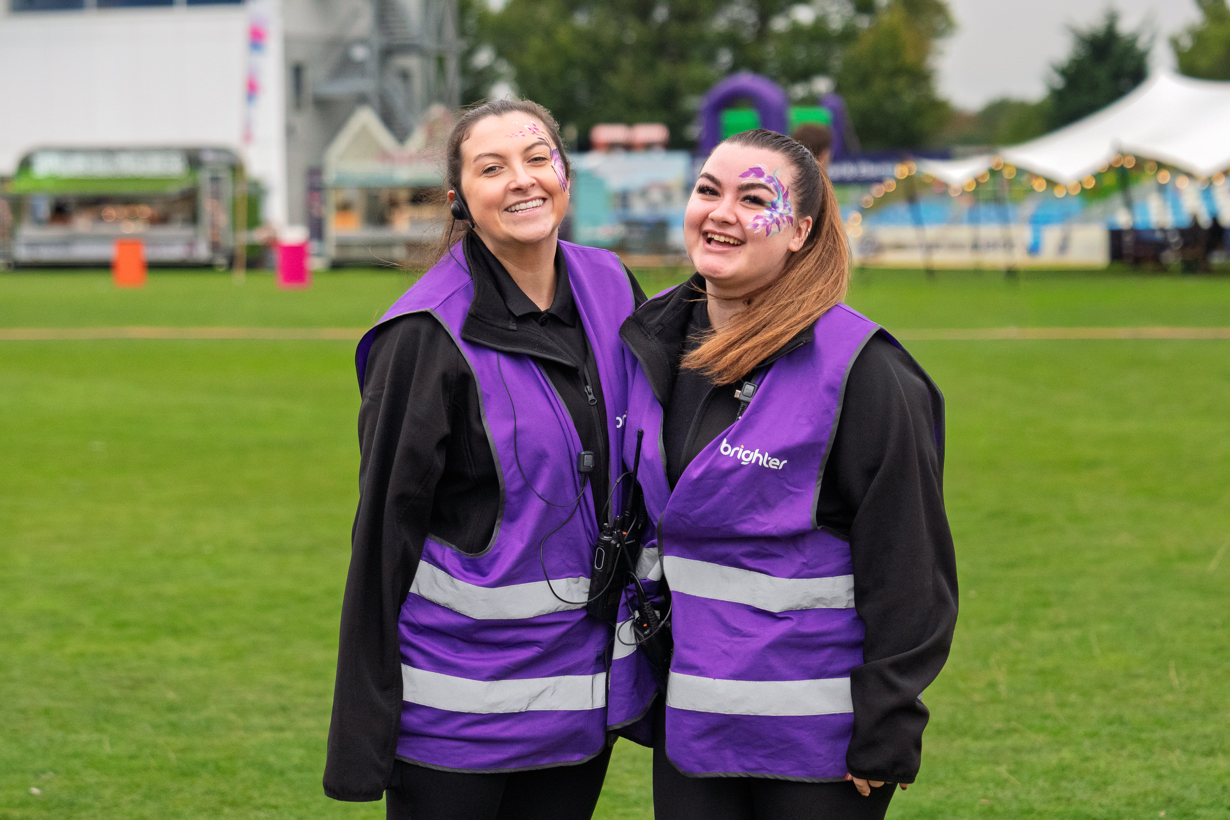 Two smiling women wearing purple safety vests and face paint standing on a grassy field at an outdoor event.