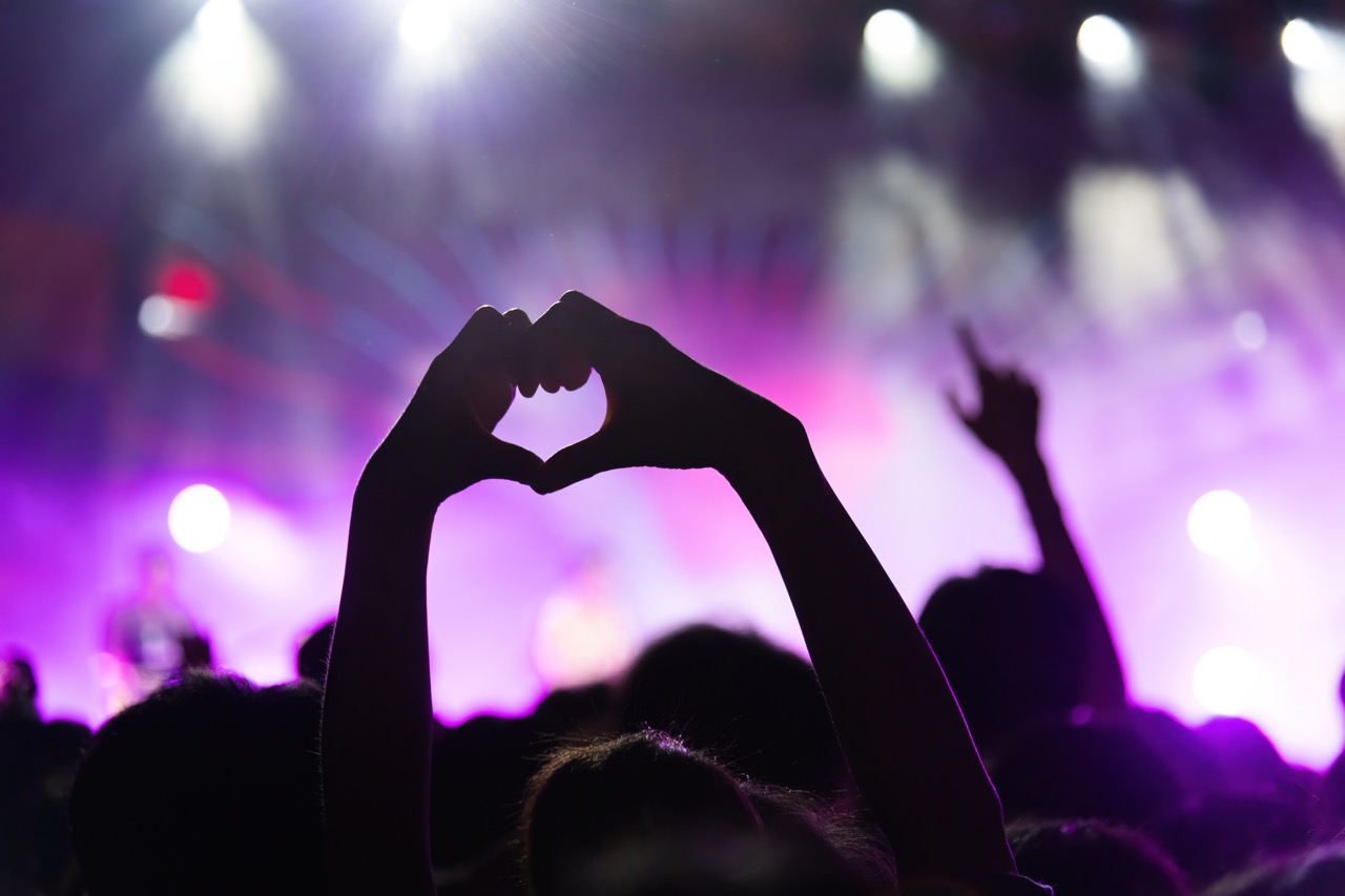Audience member forming a heart shape with hands at a concert with purple stage lights.