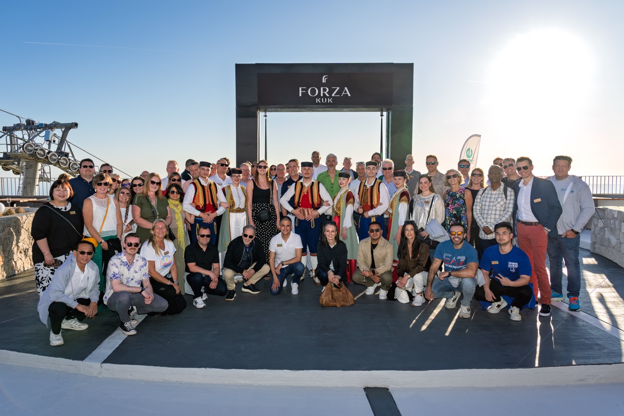 Large group of people posing outdoors in front of a Forza Kuk sign, with some wearing traditional Montenegrin attire and others in casual clothing under a clear sky.