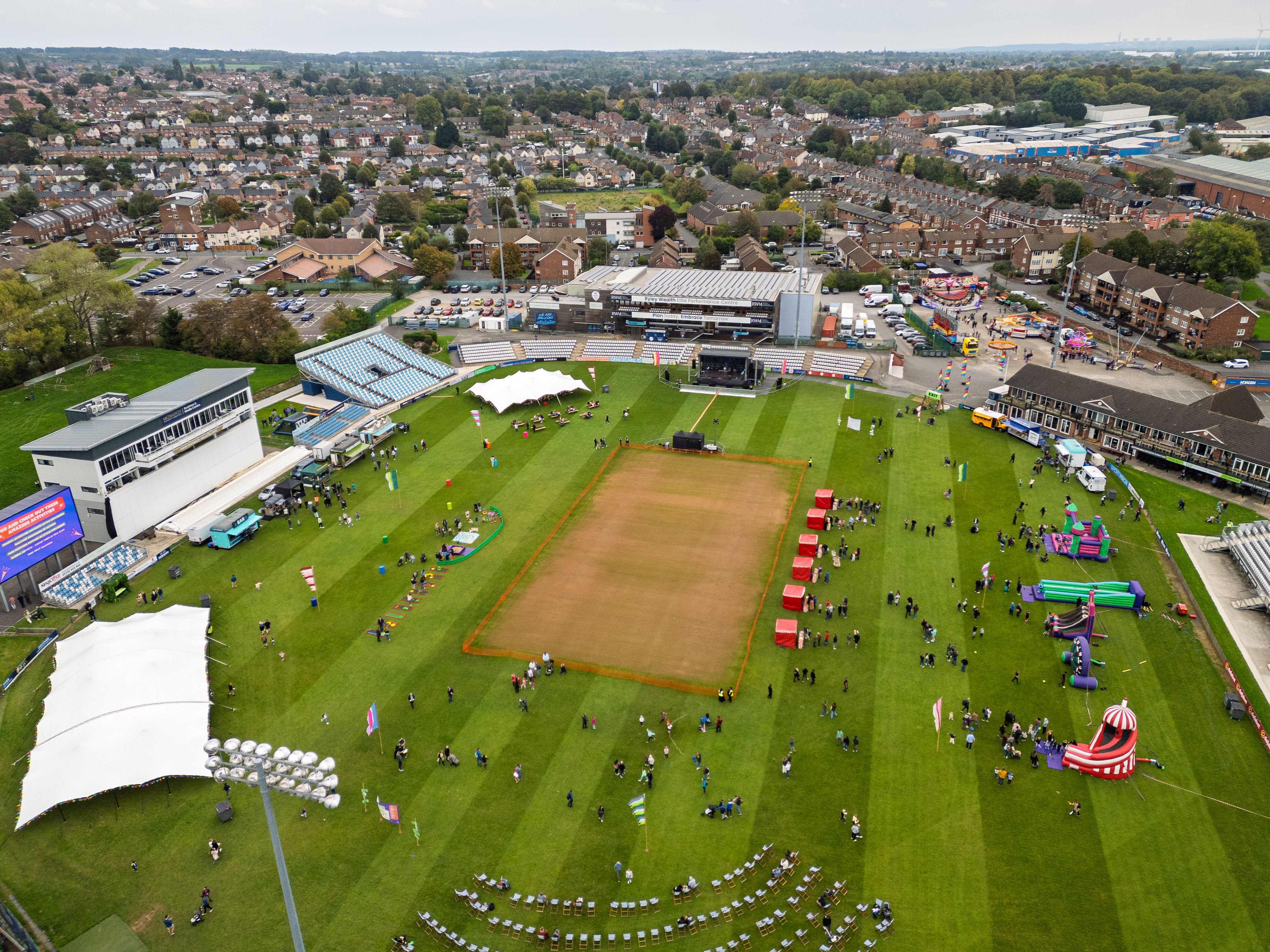 Aerial view of a sports field event with tents, inflatable attractions, scattered groups of people, and surrounding residential area.