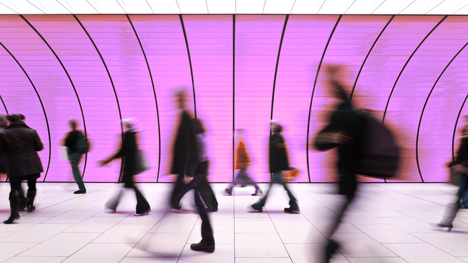 Blurred silhouettes of people walking in a tunnel with curved walls illuminated in purple light.