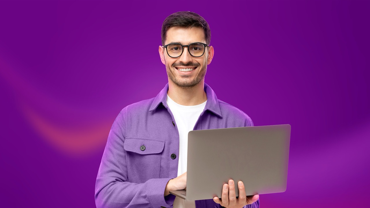 Smiling man wearing glasses and purple jacket holding an open laptop against a purple background.