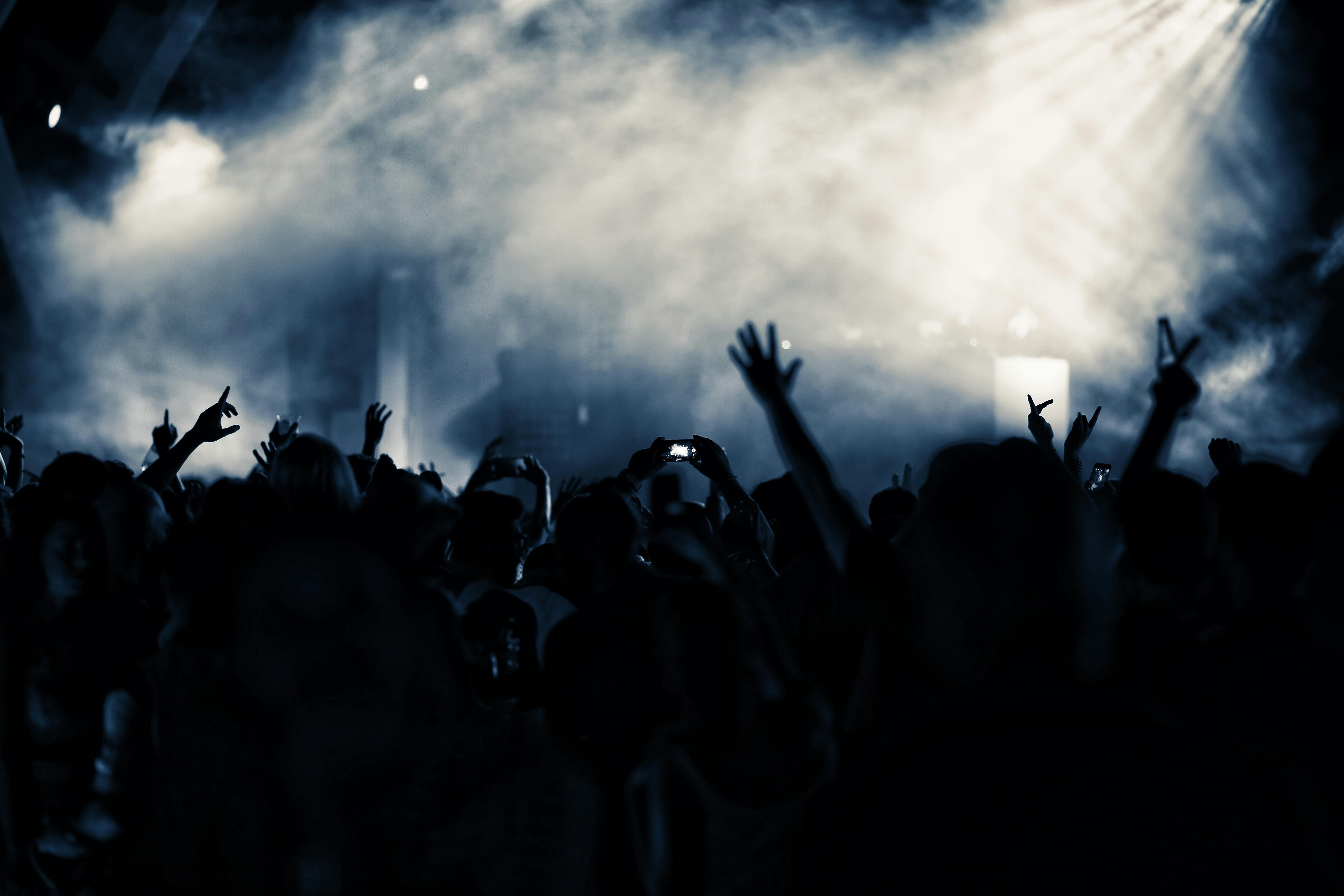 Crowd with raised hands cheering and recording at a foggy concert under bright stage lights.