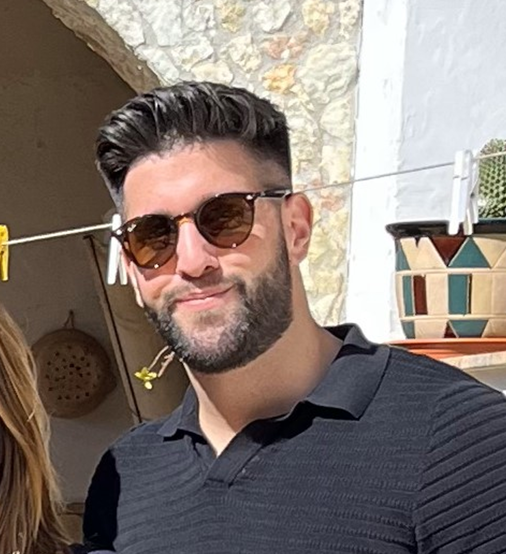 Man with dark hair and beard wearing sunglasses and a black collared shirt, standing outdoors near a stone wall and a decorative planter.