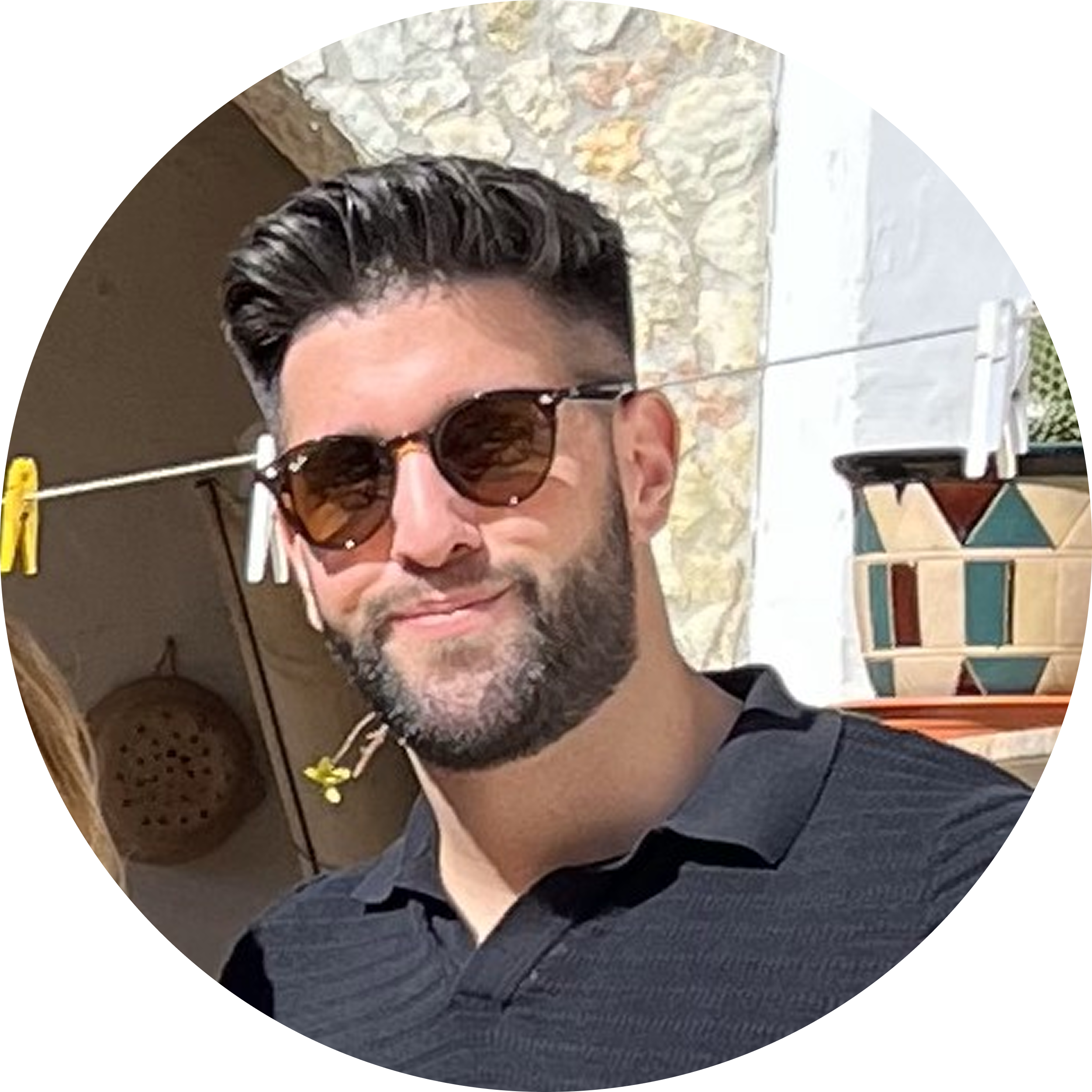 Man with dark hair and beard wearing sunglasses and a black collared shirt, standing outdoors near a stone wall and colorful pot.