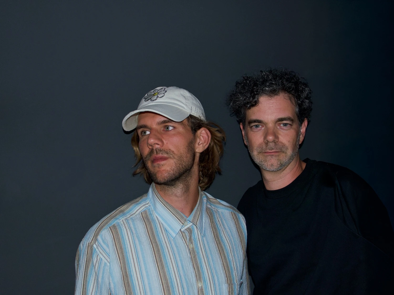 Zwei Männer stehen nebeneinander vor schwarzer Wand. Links Basti mit Cap und Blick leicht abgewandt und rechts Thomas mit dunklen Haaren und Blick zur Kamera.