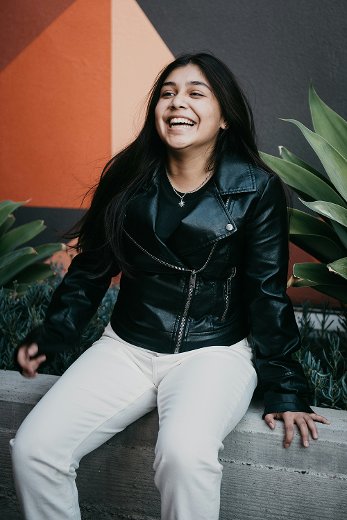 Smiling young woman with long dark hair wearing a black leather jacket and white pants sitting outdoors on a concrete ledge near green plants and a colorful geometric wall.