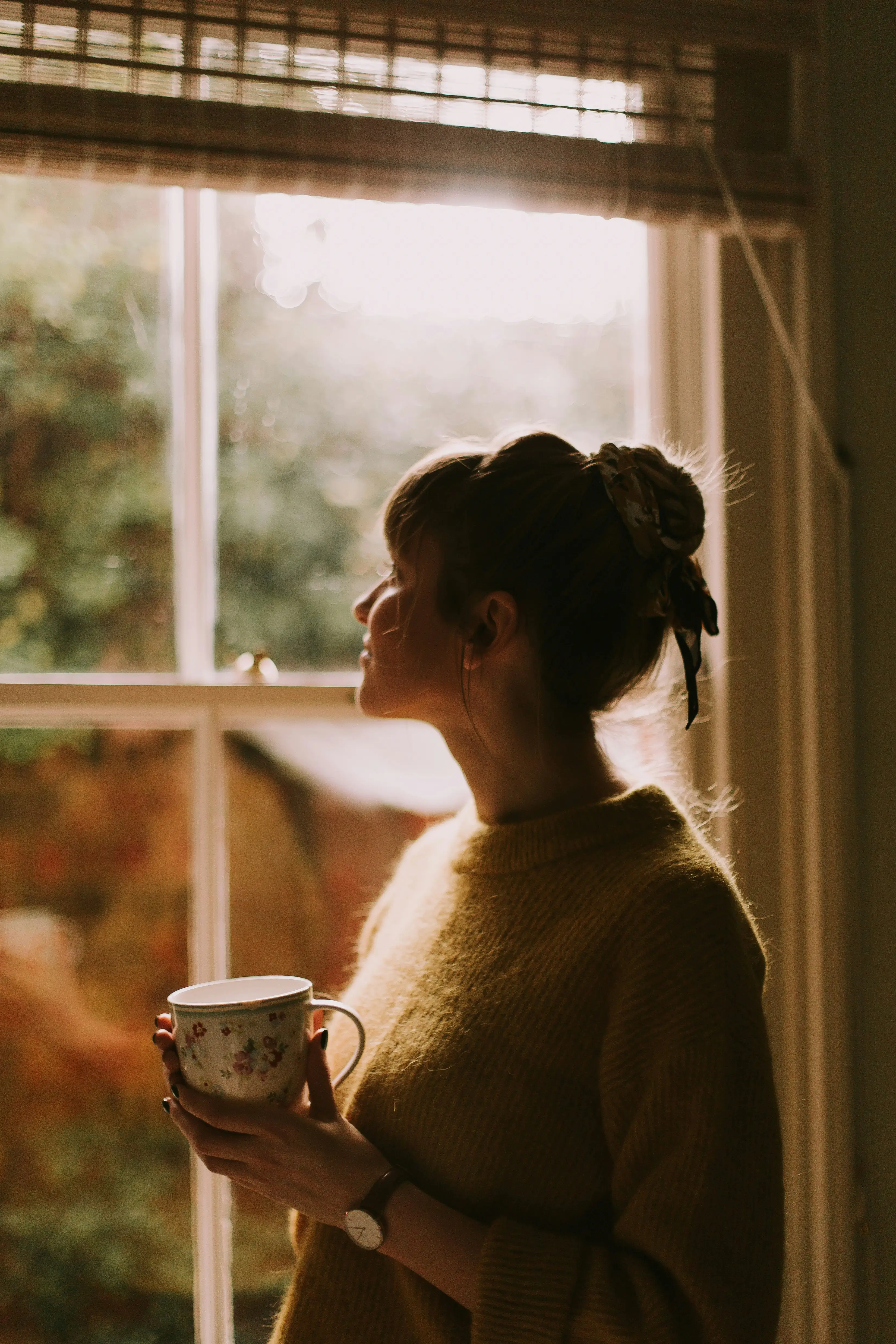 Woman in a cozy sweater holding a floral mug and looking out a sunlit window.
