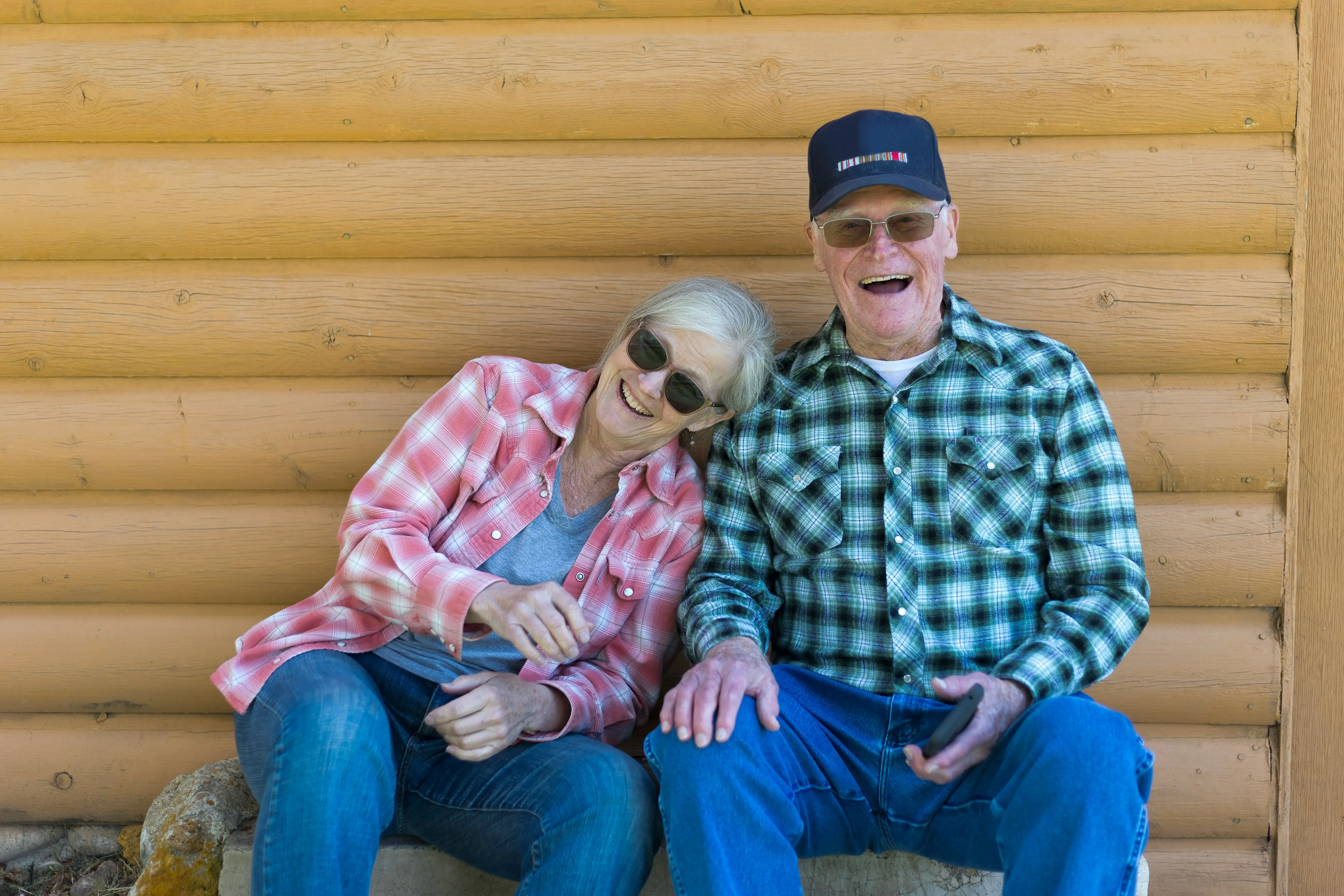 Smiling elderly couple wearing sunglasses and plaid shirts sitting against a wooden log wall.