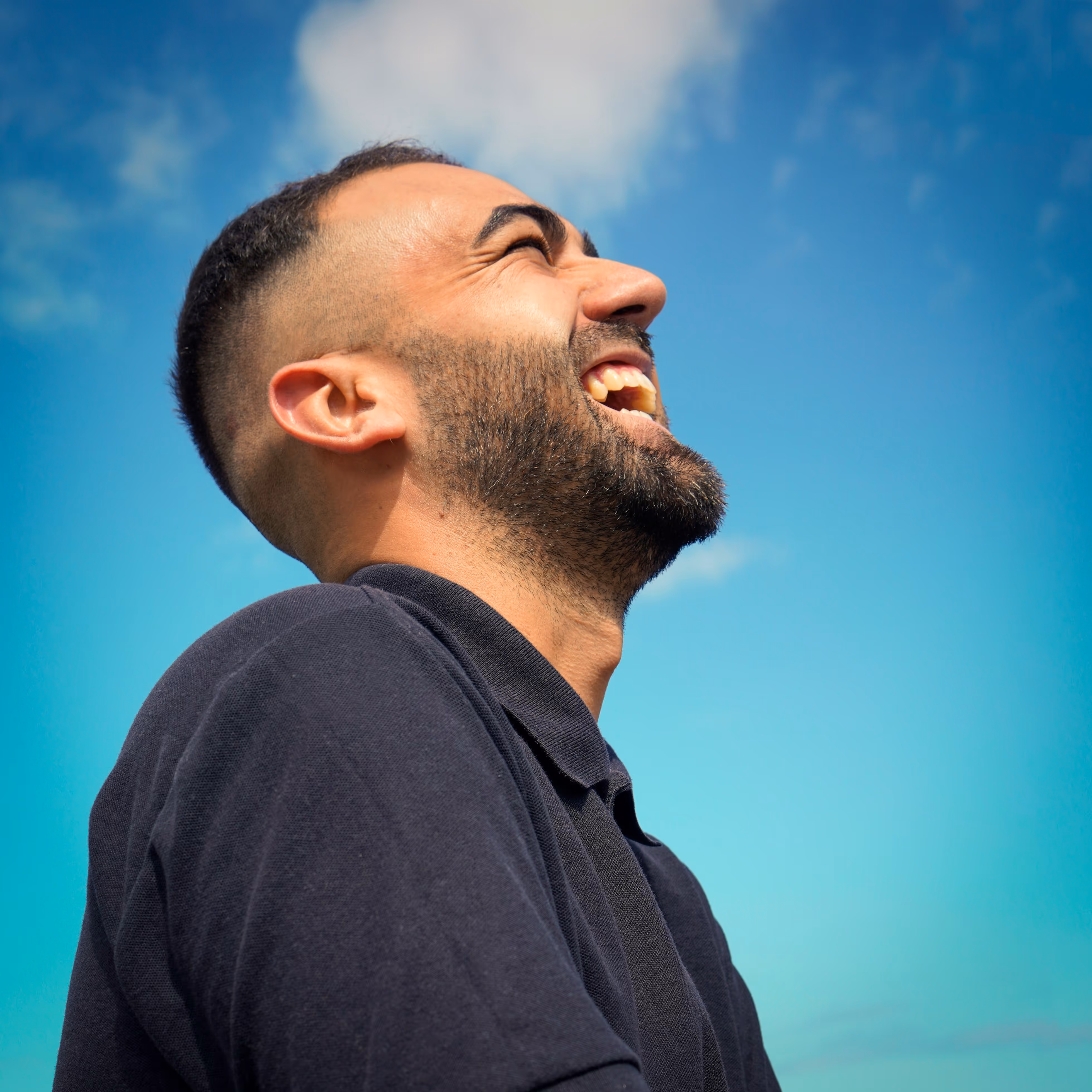 Close-up profile of a man with a beard laughing joyfully against a blue sky with clouds.