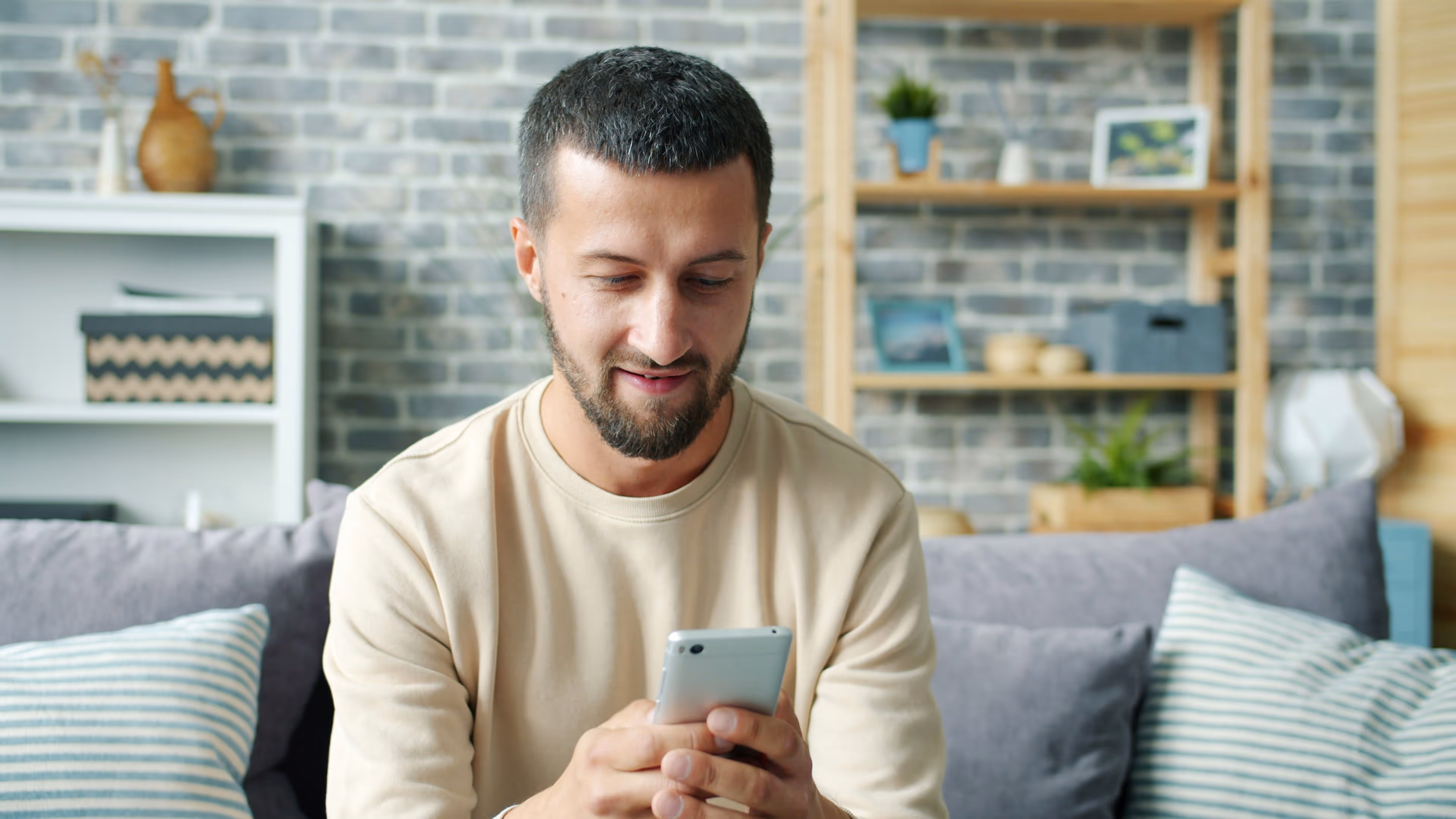 Man sitting on a couch looking at and holding a smartphone with both hands in a cozy living room.