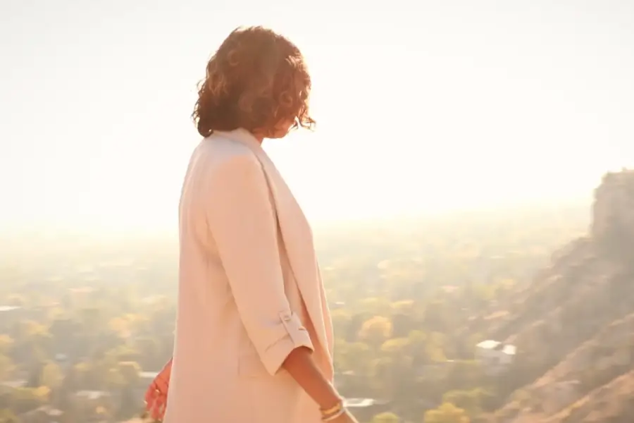 Woman with curly hair wearing a light coat standing outdoors overlooking a sunlit valley.