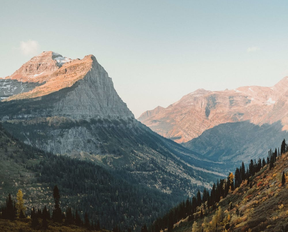 Sunlit rugged mountain peaks with forested valley and sparse autumn trees under clear sky.