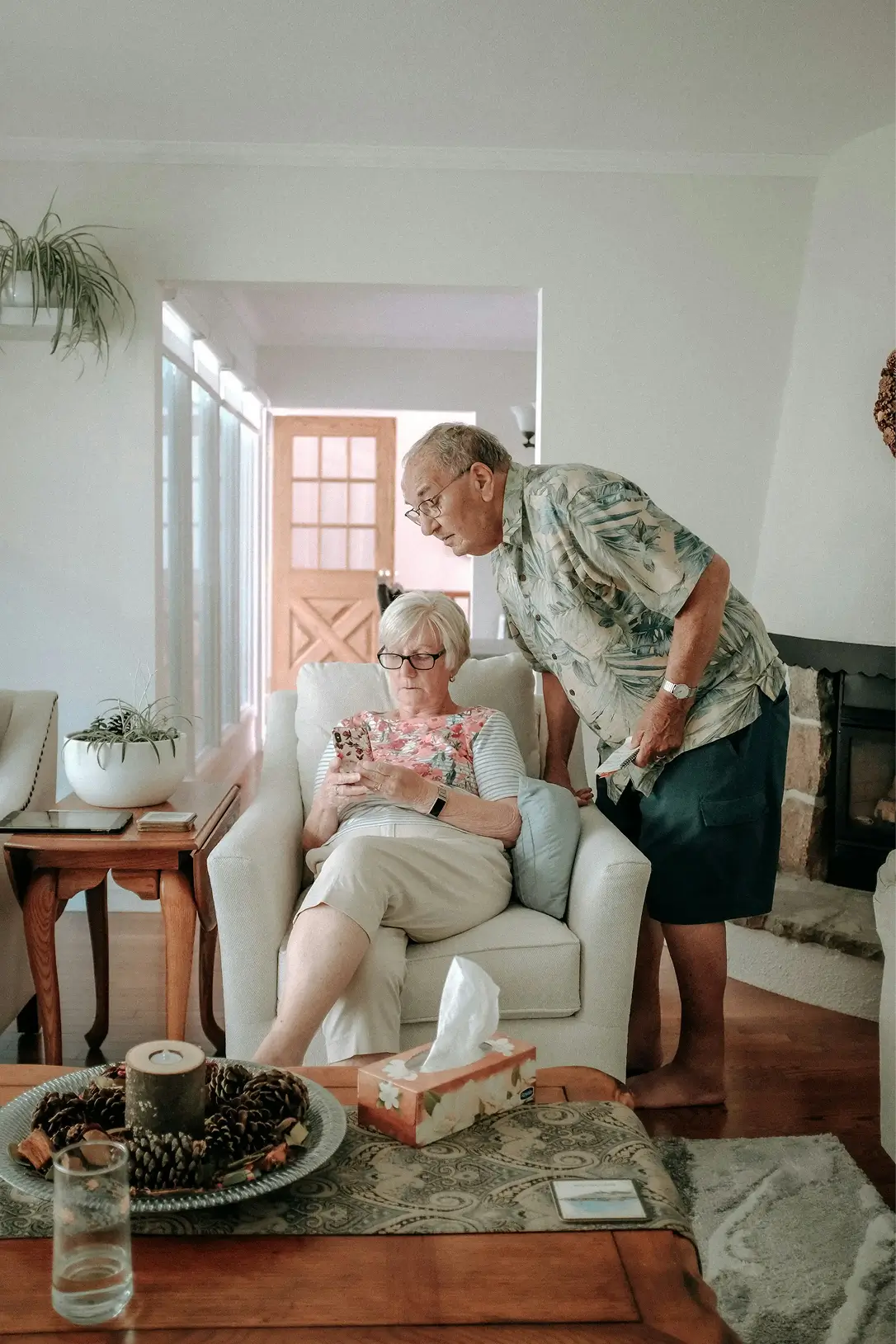 An elderly woman sitting on a beige armchair looking at her phone while an elderly man leans over beside her in a living room.