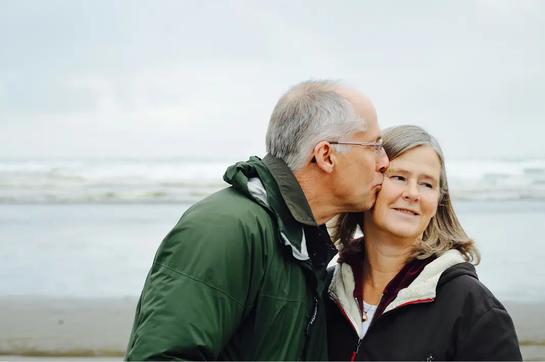 Older man in green jacket kissing a smiling woman in a black coat on the cheek at the beach.