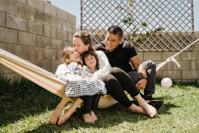 Smiling family of four sitting together on a hammock in a sunny backyard.