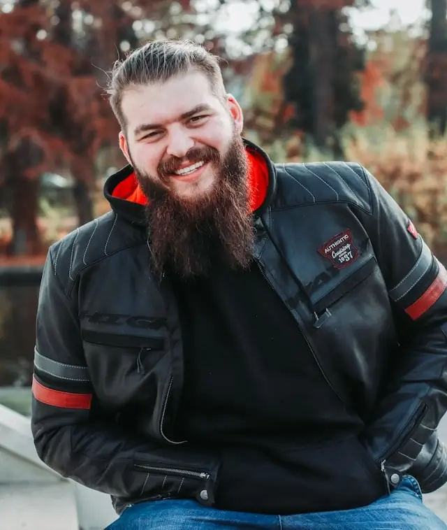 Smiling bearded man wearing a black leather jacket and blue jeans sitting outdoors with autumn foliage in the background.