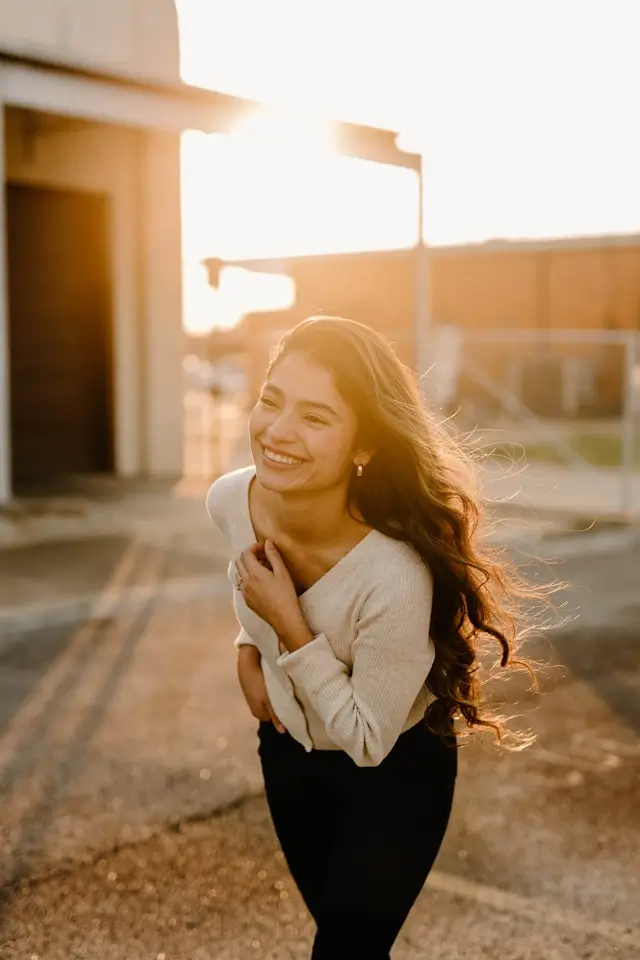 Smiling woman with long wavy hair wearing a beige sweater and black pants, leaning forward outdoors in warm sunlight.